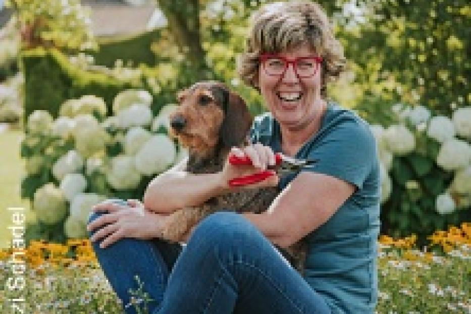 Eine Frau mit roter Brille sitzt mit einem Dackel auf einer Blumenwiese und hält eine Gartenschere.