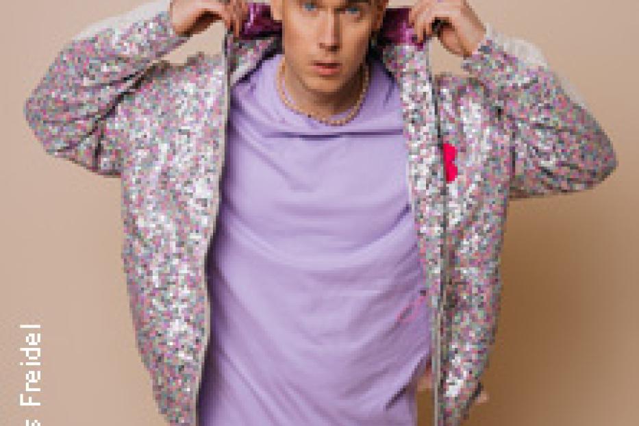 Ein junger Mann mit lila Haaren und einer glitzernden Jacke posiert vor einem beigen Hintergrund.