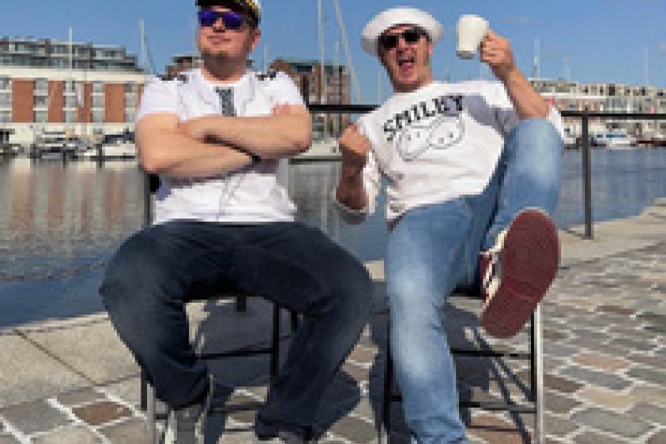 Zwei Männer in Matrosen- und Kapitänsmützen sitzen vor einem Hafen.