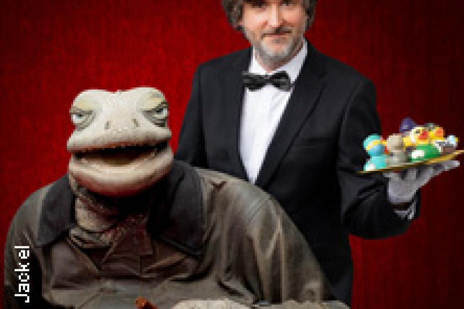 Ein Mann im Anzug präsentiert eine Echsenpuppe mit Zigarre und ein Tablett mit Süßigkeiten.