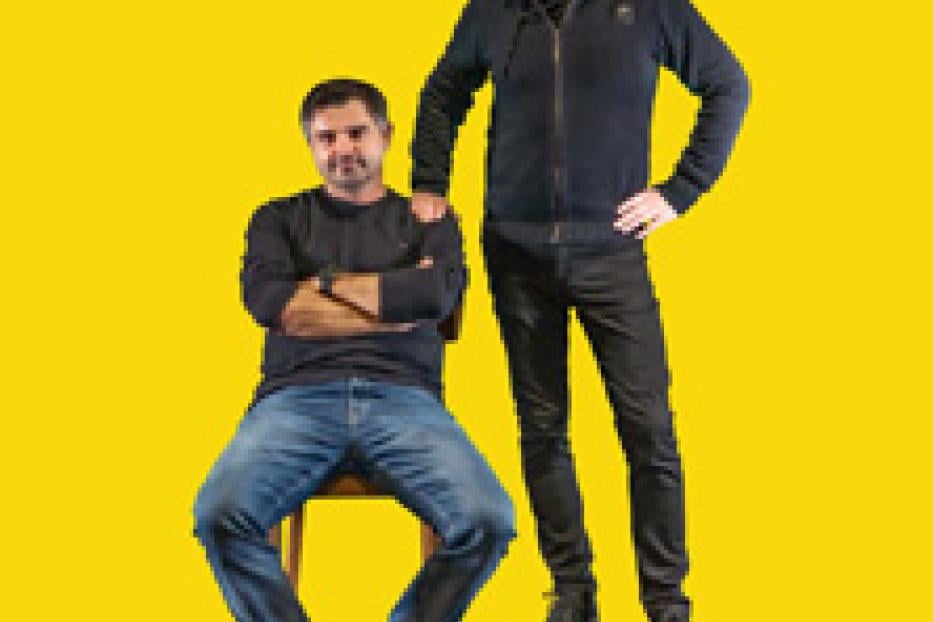 Zwei Männer posieren vor einem gelben Hintergrund, einer sitzend und einer stehend.