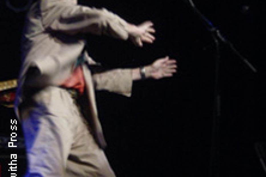 Ein Mann in einem Anzug und Hut gestikuliert auf einer Bühne.