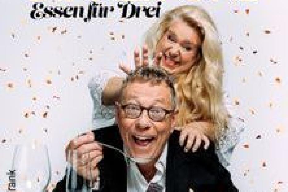 Ein Mann isst Suppe, während eine Frau hinter ihm steht und ihn neckt. Das Bild wirbt für „Dinner for One – Essen für Drei“.