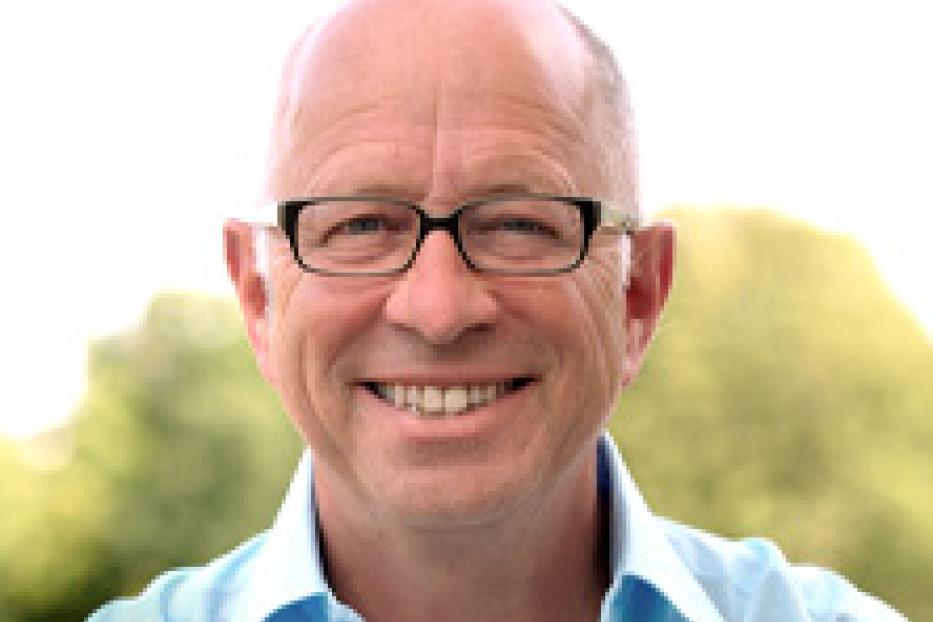 Ein lächelnder Mann mit Brille und hellblauem Hemd vor einem verschwommenen, grünen Hintergrund.