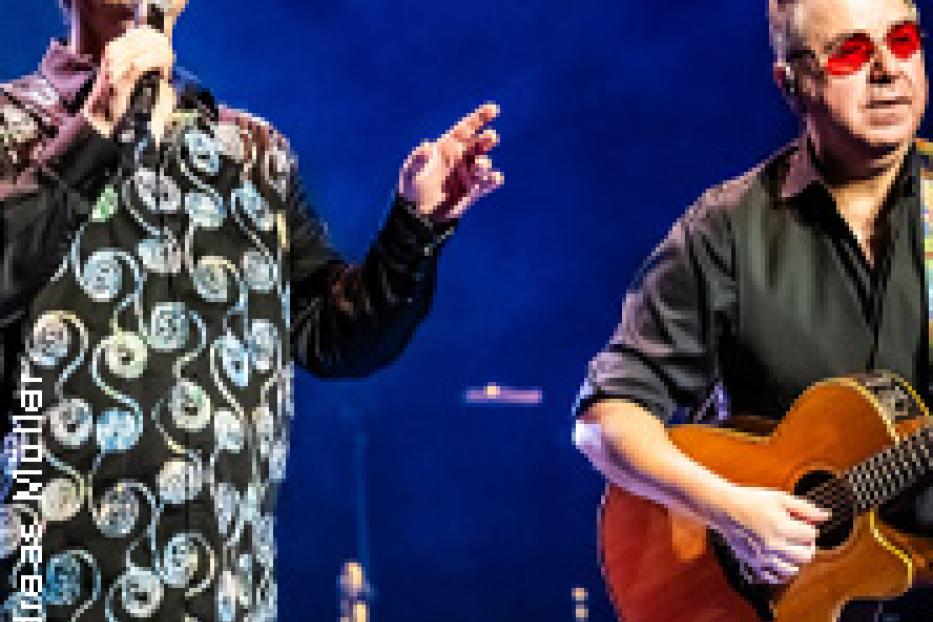 Zwei Männer stehen auf einer Bühne, einer singt ins Mikrofon, der andere spielt Gitarre.