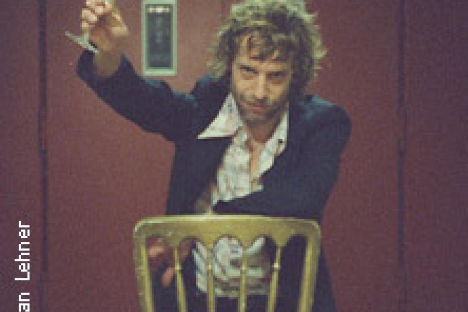 Ein Mann mit lockigem Haar sitzt auf einem goldenen Stuhl und hält ein Weinglas hoch.