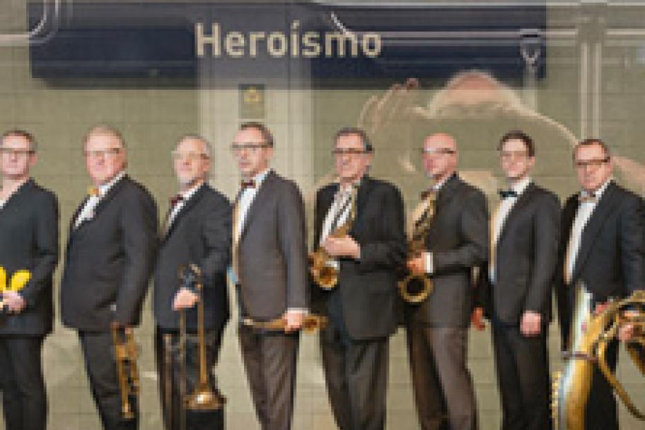 Eine Gruppe von acht Männern in Anzügen steht vor einem U-Bahn-Schild mit der Aufschrift „Heroísmo“ und hält Musikinstrumente.