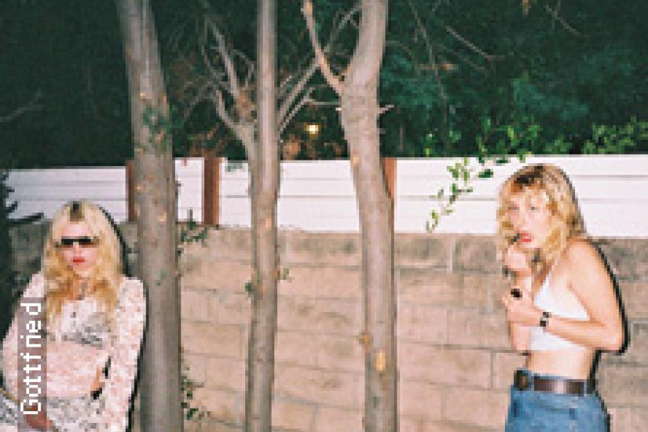 Zwei junge Frauen posieren vor einer Mauer und Bäumen, eine raucht.