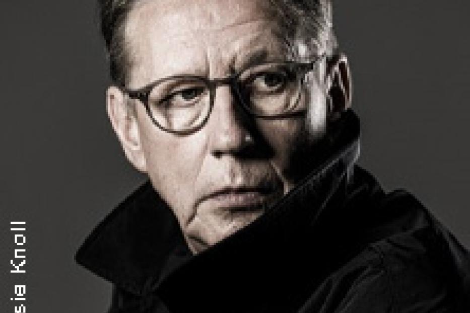 Ein Porträt von Götz Alsmann mit Brille und dunklem Hemd vor grauem Hintergrund.