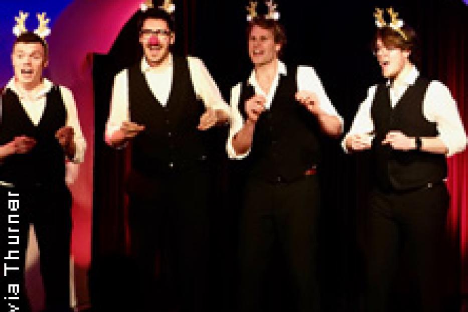 Eine Gruppe von vier Männern in Anzügen und Rentiergeweihen singt auf einer Bühne.