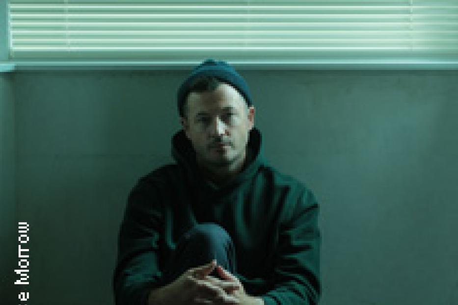 Ein Mann mit Mütze sitzt vor einem Fenster mit geschlossenen Jalousien.