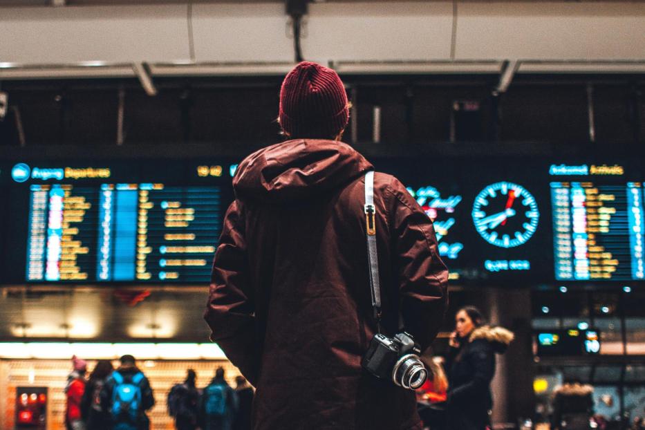 Person mit roter Jacke und Haube steht am Flughafen vor einer großen Anzeigetafel. Um ihre Schulter hängt eine Spiegelreflexkamera. 