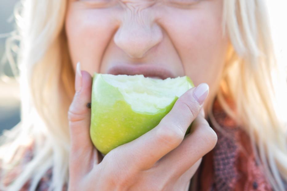 Eine blonde Frau beißt in einen grünen Apfel und verzieht dabei das Gesicht