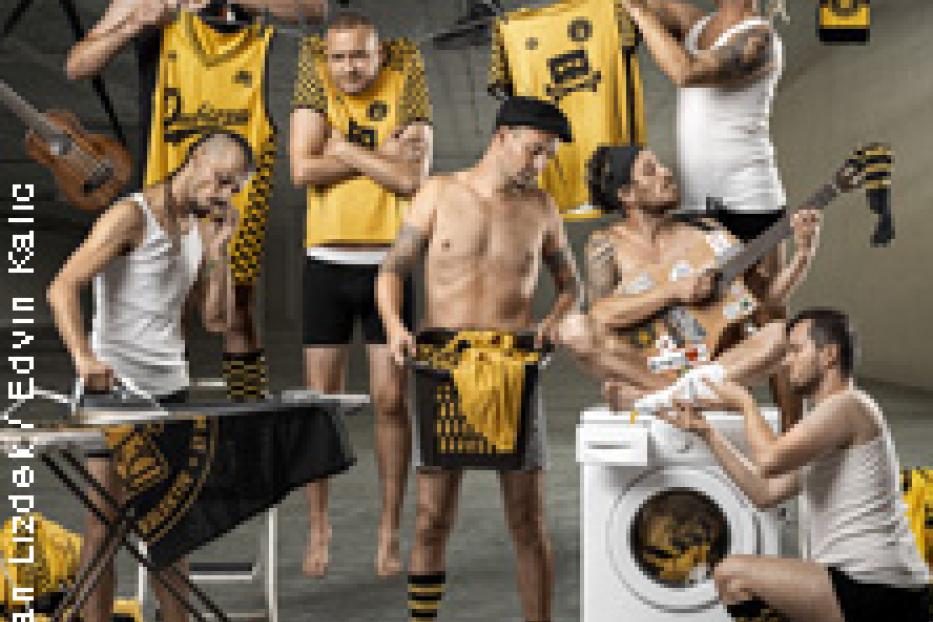 Eine Gruppe von Männern posiert mit gelb-schwarzen Kleidungsstücken und Requisiten wie Bügeleisen und Waschmaschine.