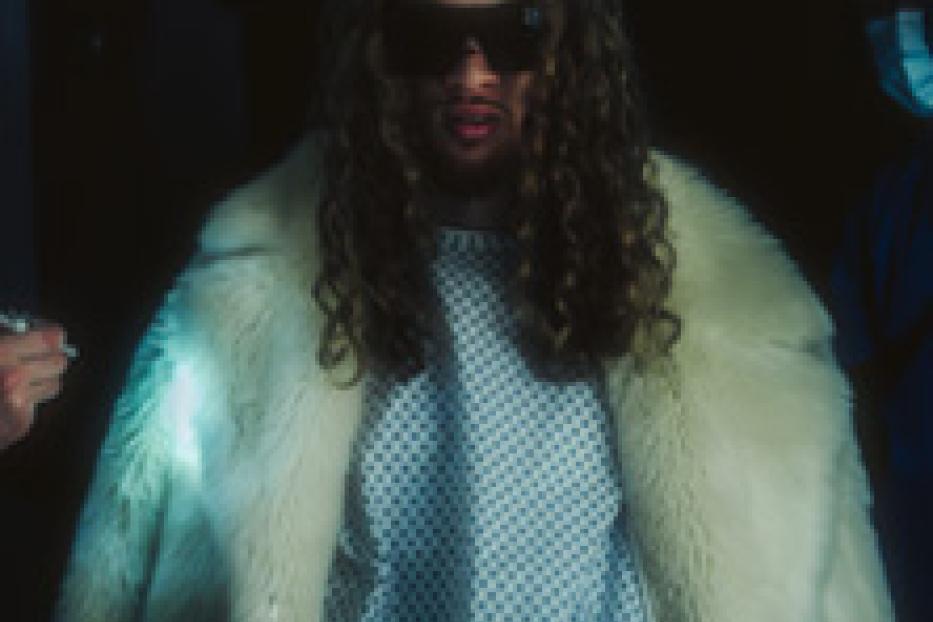 Ein Mann mit langen lockigen Haaren, Sonnenbrille und einem weißen Pelzmantel über einem Krankenhaushemd.