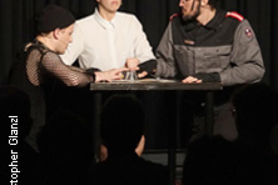 Drei Schauspieler stehen an einem Tisch während einer Theatervorstellung.