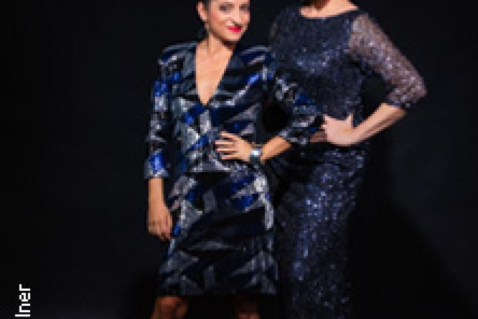 Zwei Frauen posieren in glitzernden Kleidern vor einem schwarzen Hintergrund.