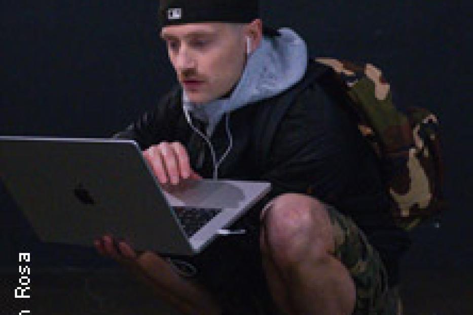 Ein Mann mit Mütze hockt und arbeitet an einem Laptop.