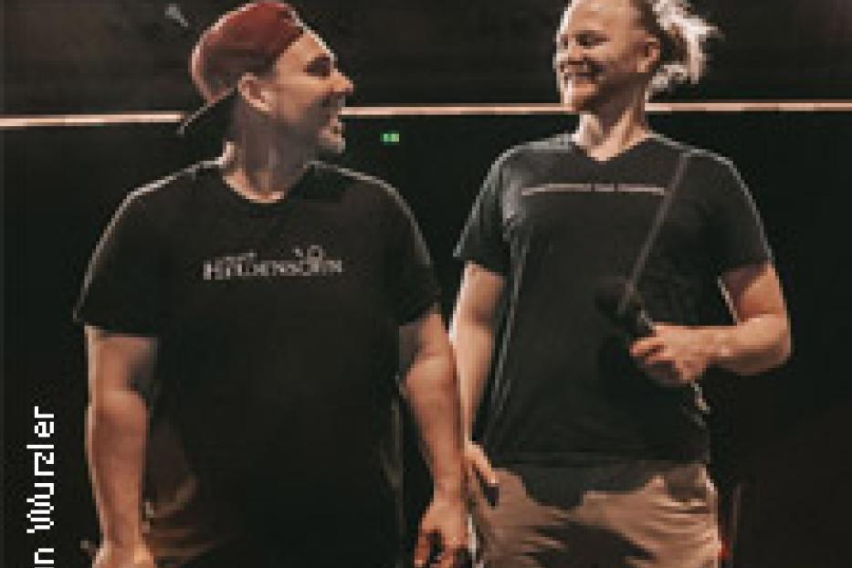 Zwei Männer stehen auf einer Bühne und lächeln sich an.