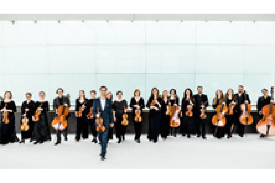 Ein Orchester steht vor einer hellen Wand und hält seine Instrumente.