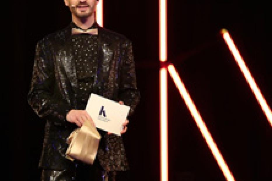 Ein Mann in einem glitzernden Anzug hält eine goldene Tasche und ein Dokument vor einem schwarzen Hintergrund.