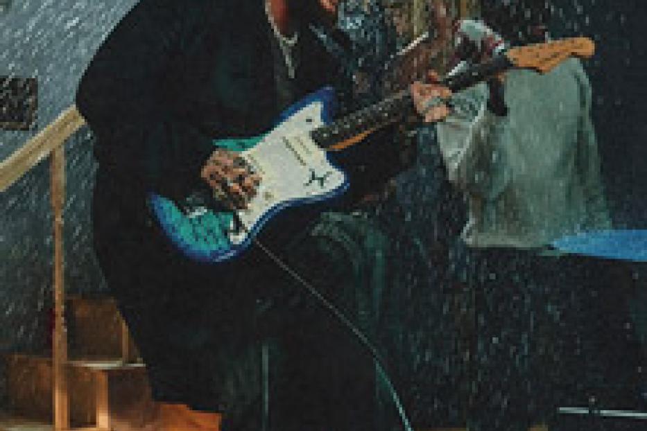 Ein Mann mit Tätowierungen spielt Gitarre, während es regnet.