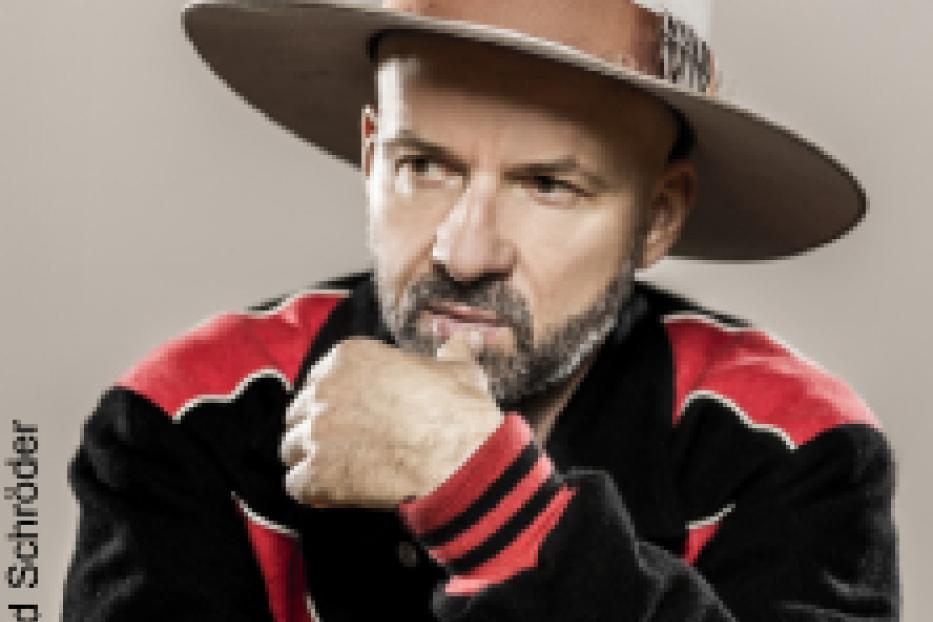 Ein Mann mit Hut und Jacke posiert vor einem hellen Hintergrund.