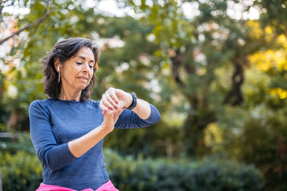 Frau um die fünfzig im Laufoutfit in einem Park schaut auf die Uhr 