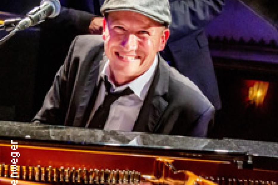 Ein lächelnder Mann mit Schiebermütze sitzt am Klavier.