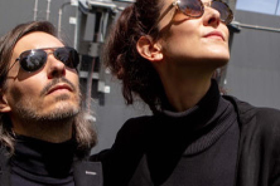 Ein Mann und eine Frau mit Sonnenbrillen stehen vor einem Gebäude und schauen nach oben.