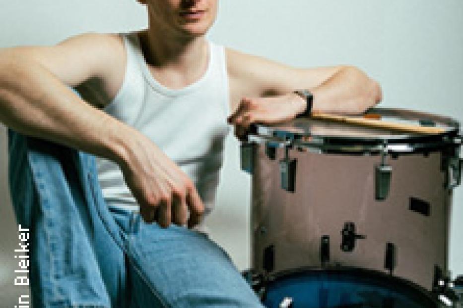 Ein junger Mann sitzt neben einem Schlagzeug und hält einen Drumstick.