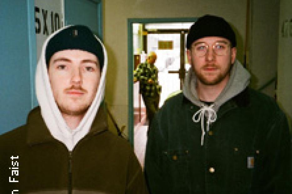 Zwei junge Männer mit Mützen stehen in einem Flur.