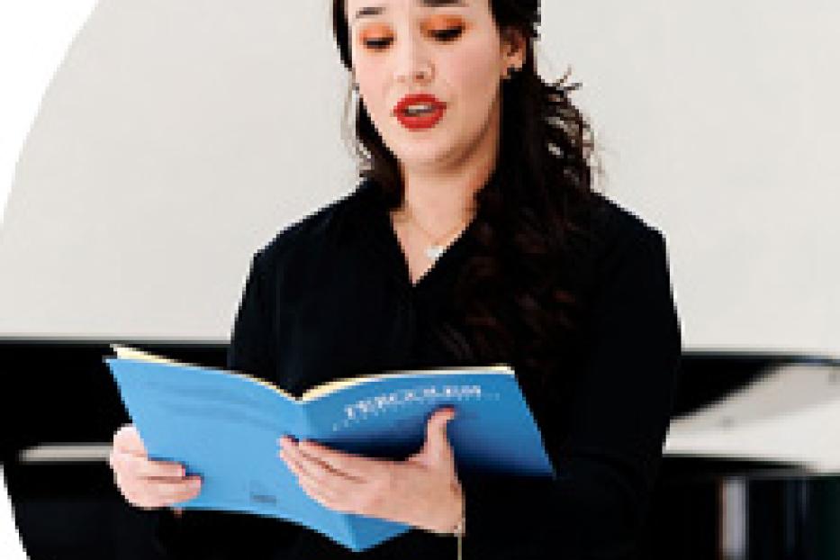 Eine junge Frau in schwarzer Kleidung singt aus einem blauen Notenbuch.