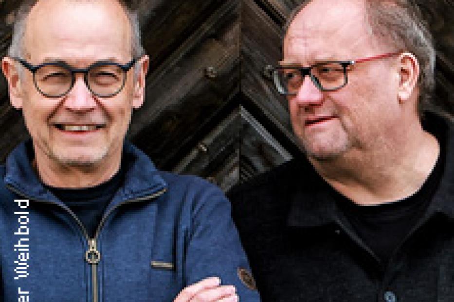 Zwei Männer mit Brillen stehen vor einer Holztür.