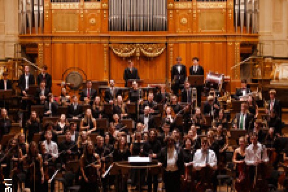 Ein Orchester spielt vor einem Publikum in einem Konzertsaal mit einer großen Orgel im Hintergrund.