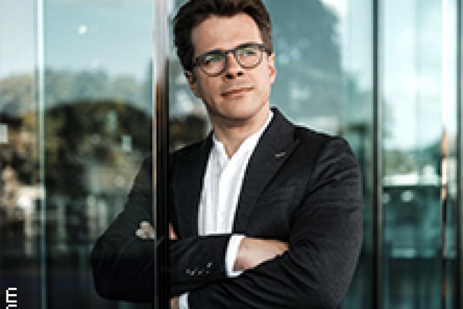 Ein Mann mit Brille und Anzug steht vor einer Glasfassade.