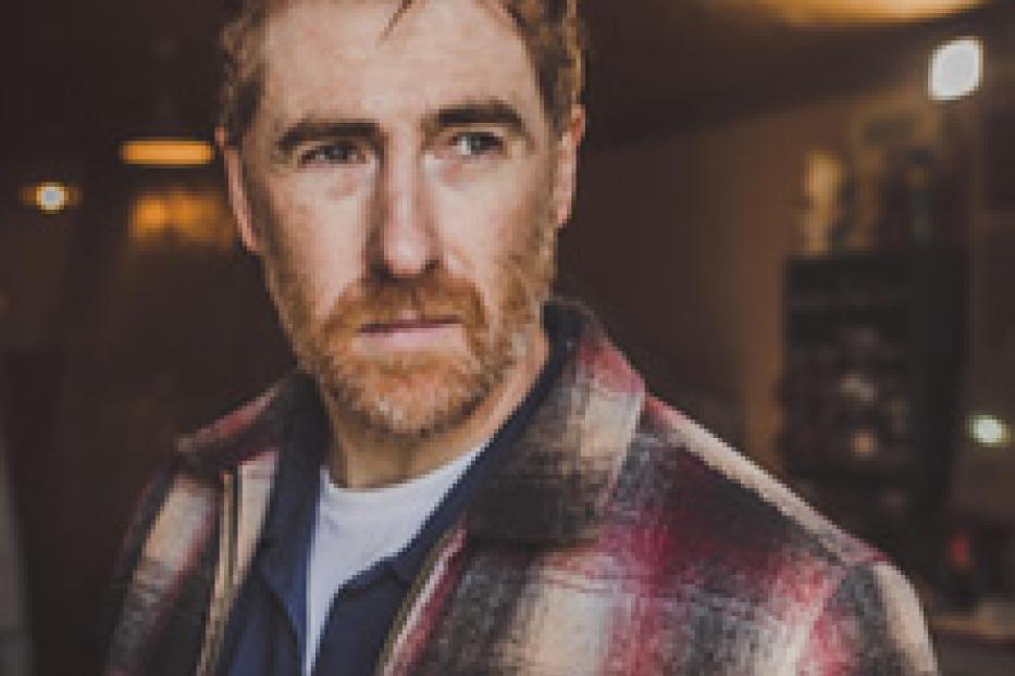 Ein Mann mit rötlichem Haar und Bart trägt ein kariertes Hemd.