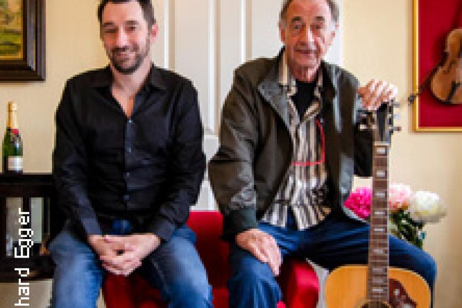 Zwei Männer sitzen vor einer Tür, einer hält eine Gitarre.