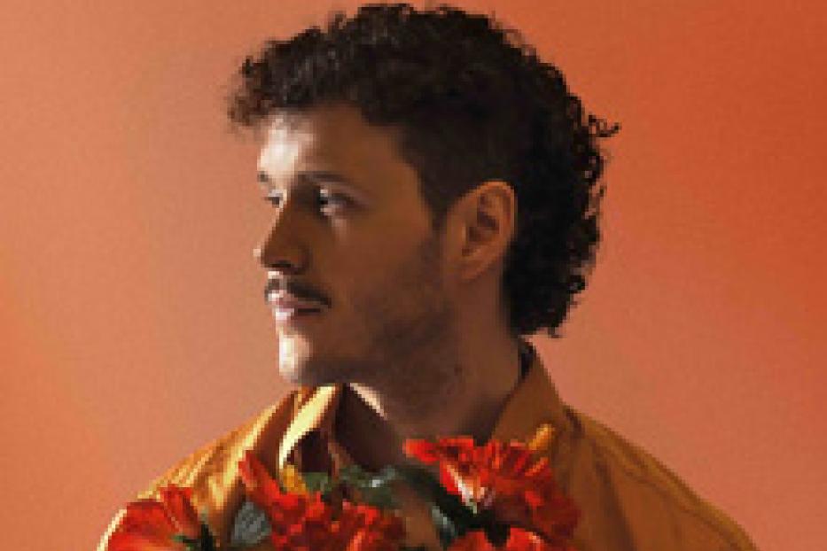 Ein Mann mit lockigem Haar und Schnurrbart hält rote Hibiskusblüten vor einem orangefarbenen Hintergrund.