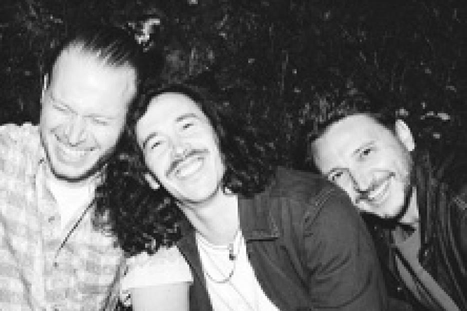 Drei Männer lachen zusammen vor einem dunklen Hintergrund.