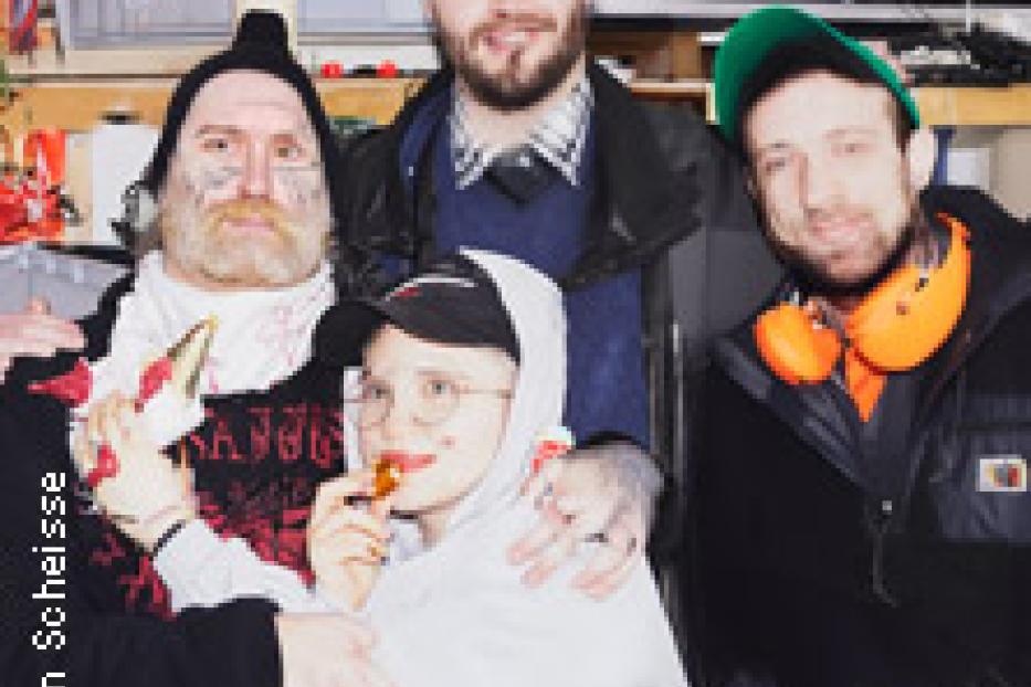 Eine Gruppe von vier Personen posiert für ein Foto, einige mit ungewöhnlichem Make-up und Hüten.