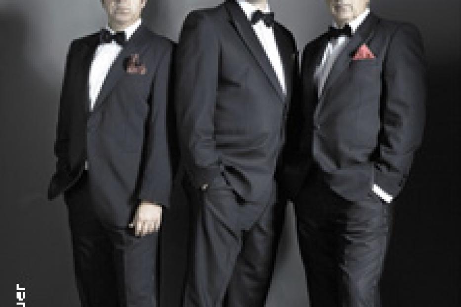 Drei Männer in Anzügen und Fliegen stehen vor einem grauen Hintergrund.