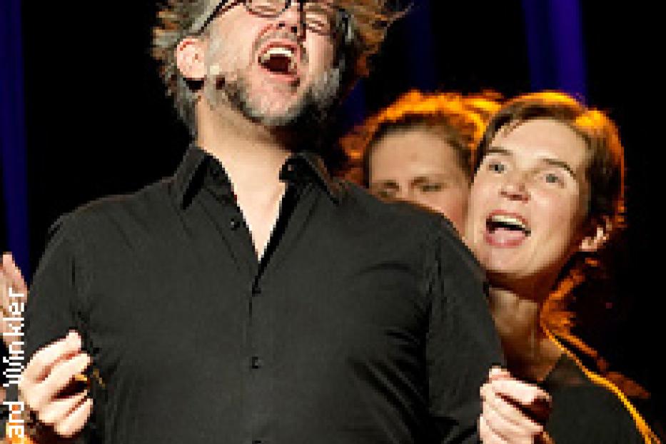 Ein Mann mit Brille und schwarzem Hemd singt mit geöffnetem Mund, begleitet von zwei Frauen.