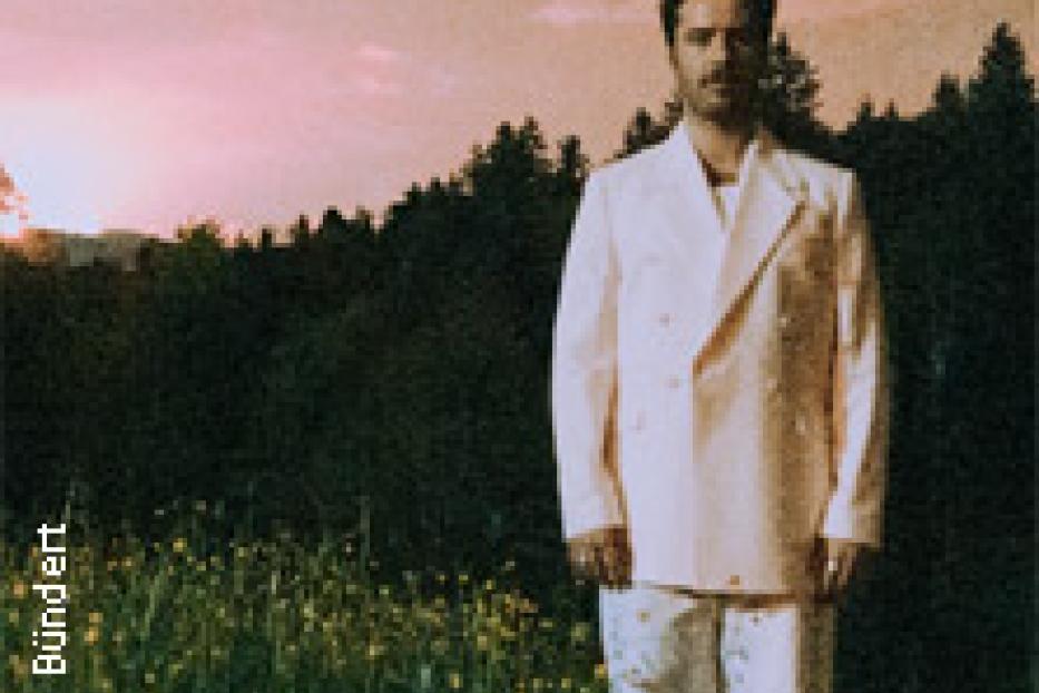 Ein Mann in einem weißen Anzug steht auf einer Wiese vor einem Wald.