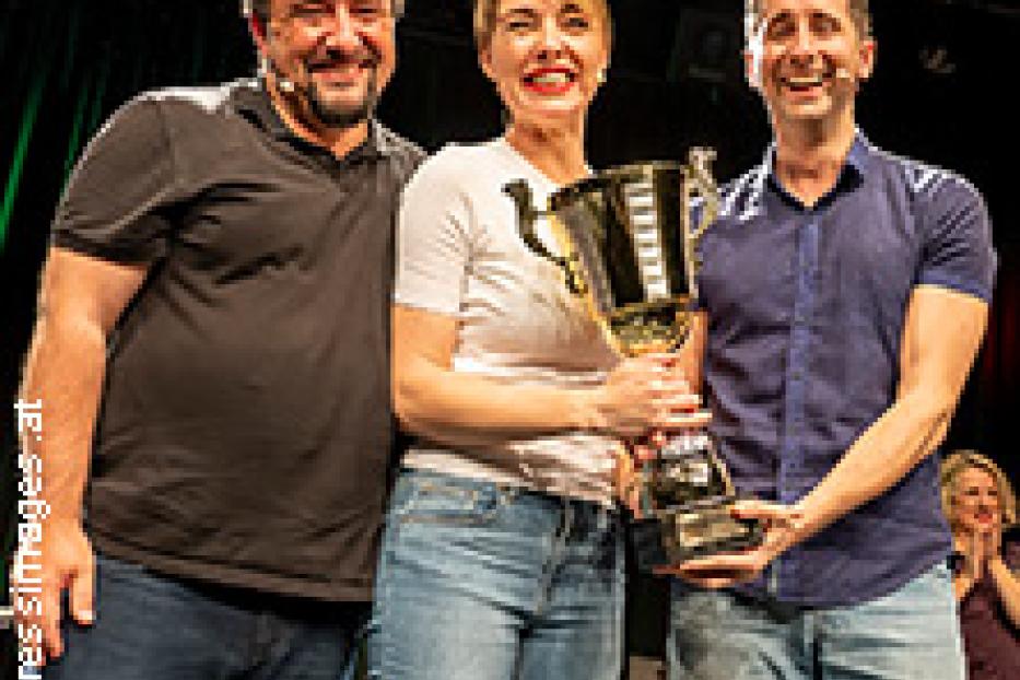 Drei Personen halten gemeinsam einen großen Pokal auf einer Bühne.