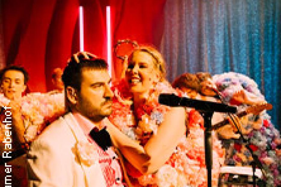 Ein Mann und eine Frau, beide mit Blumen geschmückt, stehen auf einer Bühne vor einem Mikrofon.