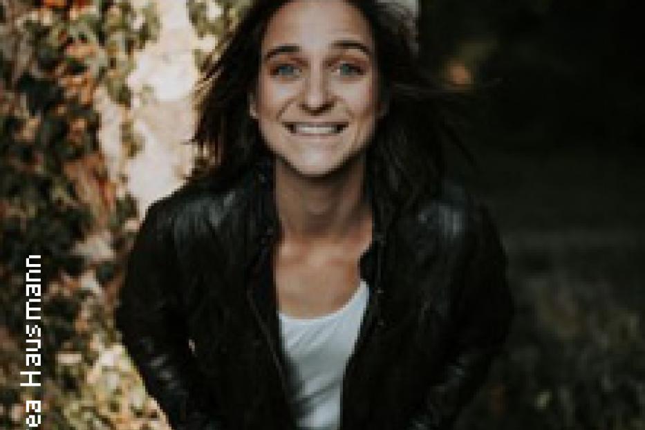 Eine lächelnde Frau in Lederjacke steht vor einer mit Efeu bewachsenen Wand.