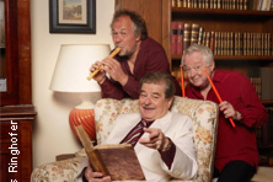 Drei ältere Herren posieren in einem Raum mit Büchern, einer spielt Flöte, einer liest ein Buch.