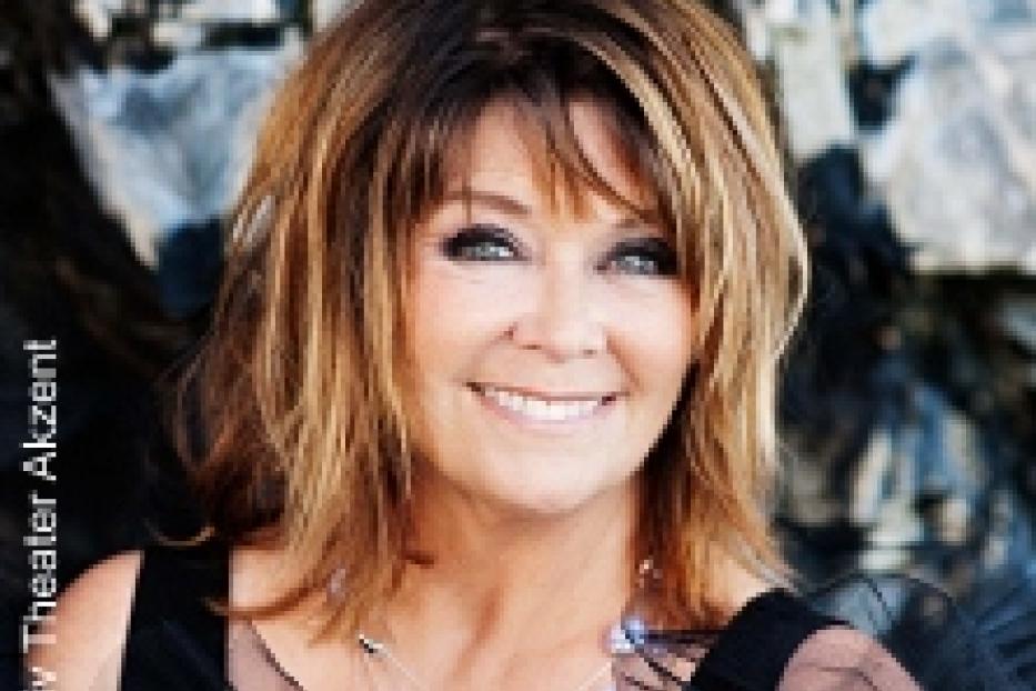 Eine lächelnde Frau mit braunen Haaren und einer Halskette vor einem Felsen.