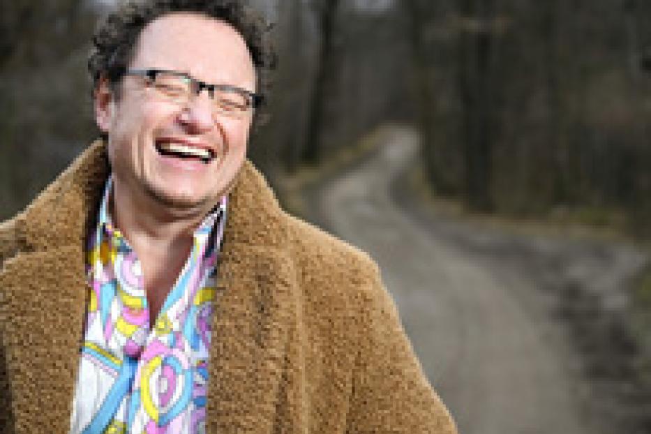 Ein lachender Mann mit Brille und braunem Mantel steht auf einem Waldweg.